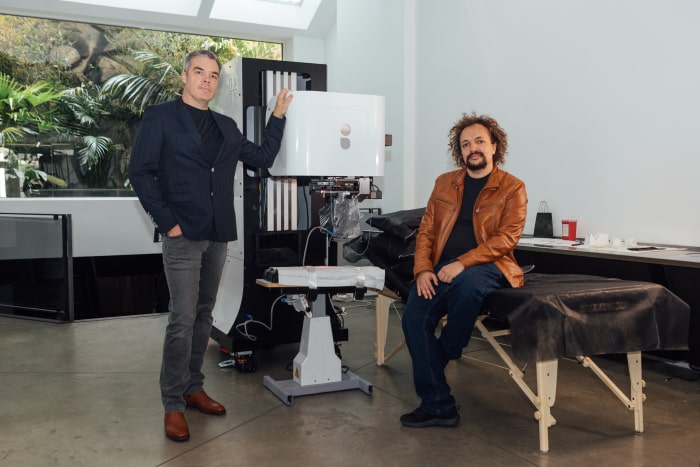 Two men standing by a tattoo machine.