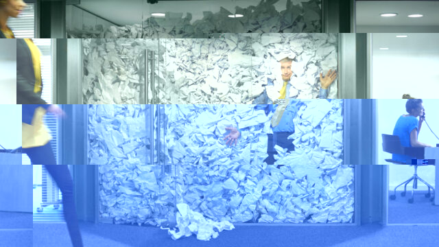 A businessman is trapped in his glass office by a surplus of discarded ideas on paper . His colleague in the next office is working more efficiently and is oblivious to him being trapped , as is a passing female office worker